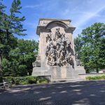 Baroque stone monument set in a park with lush green trees under a blue sky.