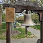 Bronze bell mounted on a wooden stand outdoors with green lawn and trees in the background.