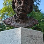 Bronze bust of Albert Einstein on a granite pedestal outdoors under trees.