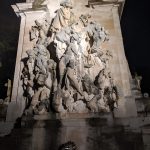 Baroque stone sculpture depicting allegorical figures on a grand architectural facade at night.