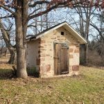 Small, rustic stone and adobe structure in a wooded, autumnal landscape.