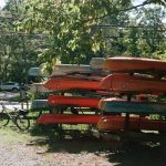 Stack of orange and blue canoes on a trailer near trees and a dirt road.