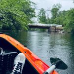 Orange kayak on a river with a wooden bridge spanning the water under green trees.