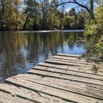 Wooden dock extends over calm river with trees lining the banks under a blue sky.