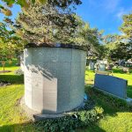 Stone mausoleum in a sunny, green cemetery setting surrounded by trees.
