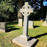 Stone Celtic cross grave marker in a grassy cemetery under large trees.