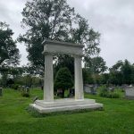 Stone mausoleum structure in a grassy, historic cemetery under an overcast sky.