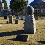 Sphere on stone monument in a grassy cemetery with headstones and trees.