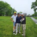 Three men stand on a green lawn in front of a building under an overcast sky.