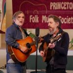 Two folk musicians perform on stage in front of a Princeton Folk Music Society banner.