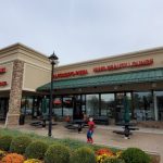 Strip mall with various storefronts, including a health food store and pizza place, on an overcast day.