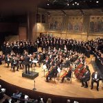 Orchestra and choir perform on a large stage in an ornate concert hall.