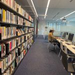Library interior with tall bookshelves lining a hallway and a person working at a desk.