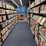 Bookstore aisle with tall shelves packed with books, leading toward a glass partition.