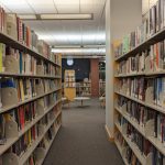 Library aisle with tall bookshelves lining a carpeted corridor, leading to a central reading area.