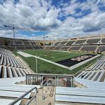 Stadium view with green field, grandstands, and construction cranes under a partly cloudy sky.