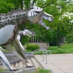Large, stylized metal sculpture of a fierce mythical creature in a park setting.
