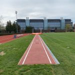 Outdoor running track with a straightaway leading toward a modern glass and steel building under an overcast sky.
