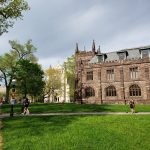Collegiate Gothic building with green lawn and trees under a cloudy sky.