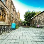 Stone courtyard between historic buildings with bicycles parked and greenery under a blue sky.