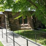 Stone building entrance with ramp and railing under leafy green trees.