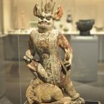Ancient ceramic deity figure with horns, seated on an animal base in a museum display.