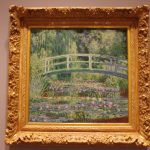 Oil painting of a lush green landscape with a white arched bridge over a flower-filled stream.