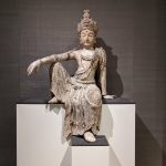 Ancient stone statue of a seated Buddha figure displayed on a modern pedestal.