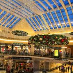 Bright, modern indoor shopping mall atrium with a glass ceiling and festive holiday decorations.