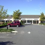 Strip mall storefronts with parking lot under a clear blue sky.