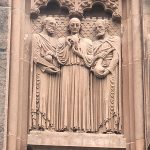 Stone relief sculpture of three robed male figures beneath an arched architectural detail.