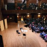 Orchestra rehearsal in a large, tiered concert hall with musicians seated on the floor.
