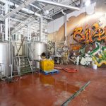 Industrial brewery interior with stainless steel tanks and vibrant graffiti on the brick wall.