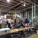 Industrial interior with high ceiling, featuring stainless steel tanks and a large communal dining table.