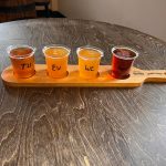 Four small cups of amber-colored drinks labeled "Th," "Ew," "We," and "Oot" on a wooden tray.