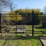Black metal fence enclosing a grassy yard with trees under a clear blue sky.
