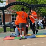 Group of children in orange shirts practicing gymnastics under an outdoor archway.