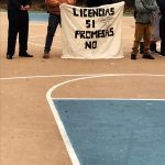 Protest sign reading "Licencias SI Promesas NO" held by people on a basketball court.