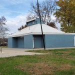 Blue, single-story building with a white roof next to a chain-link fence and grassy area.