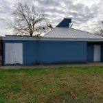 Small blue utility building with white trim, set on a grassy lawn next to a black fence.
