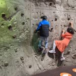 Children climb a textured, artificial rock wall in an indoor climbing gym setting.