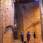 Indoor climbing gym with climbers on various walls; people watch below.