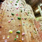 Indoor climbing wall with numerous colorful holds and ropes against a textured, tan surface.