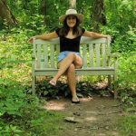 Woman in hat sits on a park bench surrounded by lush green forest trees.