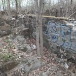 Ruined stone wall with graffiti, surrounded by trees and debris in an overgrown area.