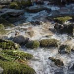 Rocky stream with moss-covered stones flowing over clear, cold water.