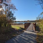 Wooden bridge crosses a calm lake under a bright blue sky, framed by autumn trees.