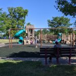 Children play on playground equipment under sunny skies with trees in the background.