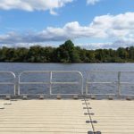 Dockside view across a calm lake with a dense line of trees under a partly cloudy sky.