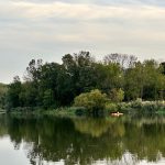 Green trees line the shore of a calm lake under a light sky.